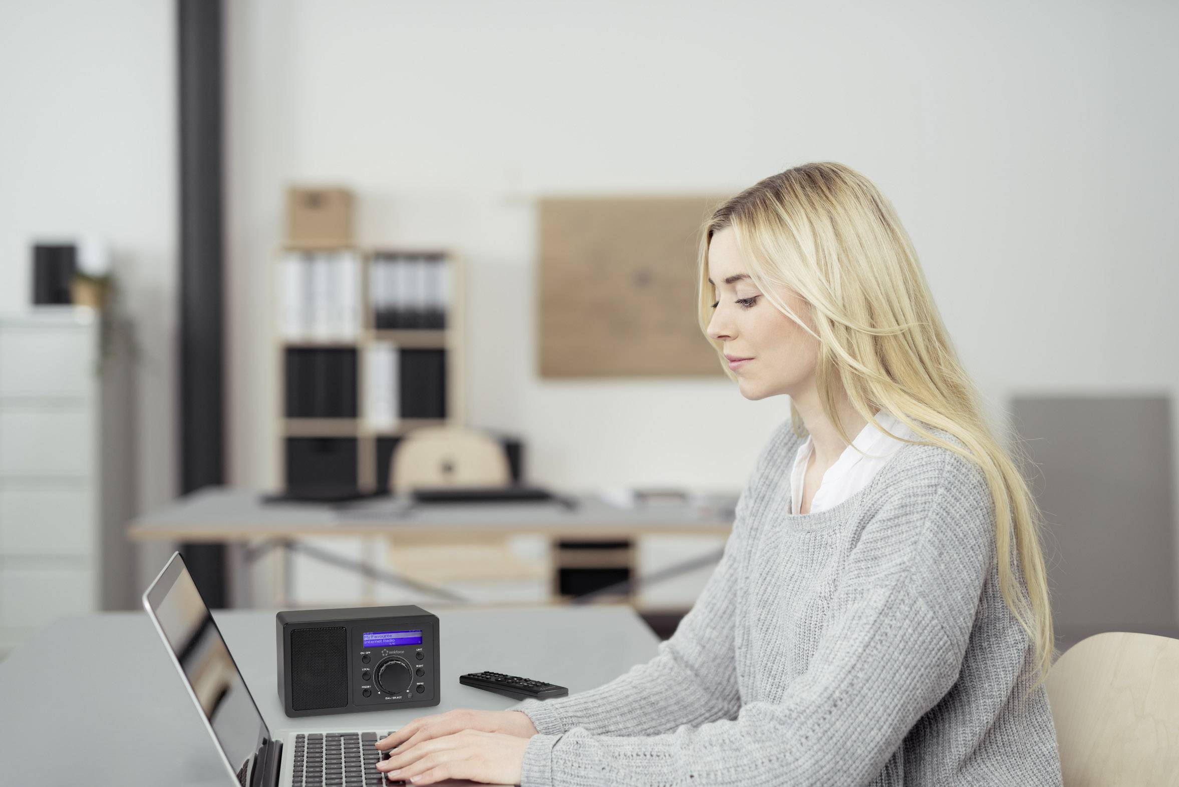 Une femme aux cheveux blonds est assise à un bureau et travaille avec concentration sur un ordinateur portable. À côté se trouve un petit haut-parleur noir.