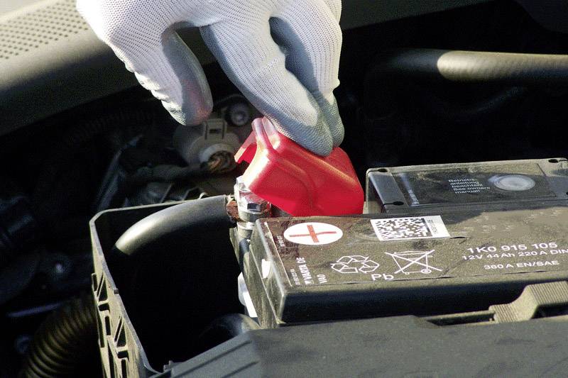Personne inspectant une borne de batterie rouge sur une batterie de voiture dans le compartiment moteur.