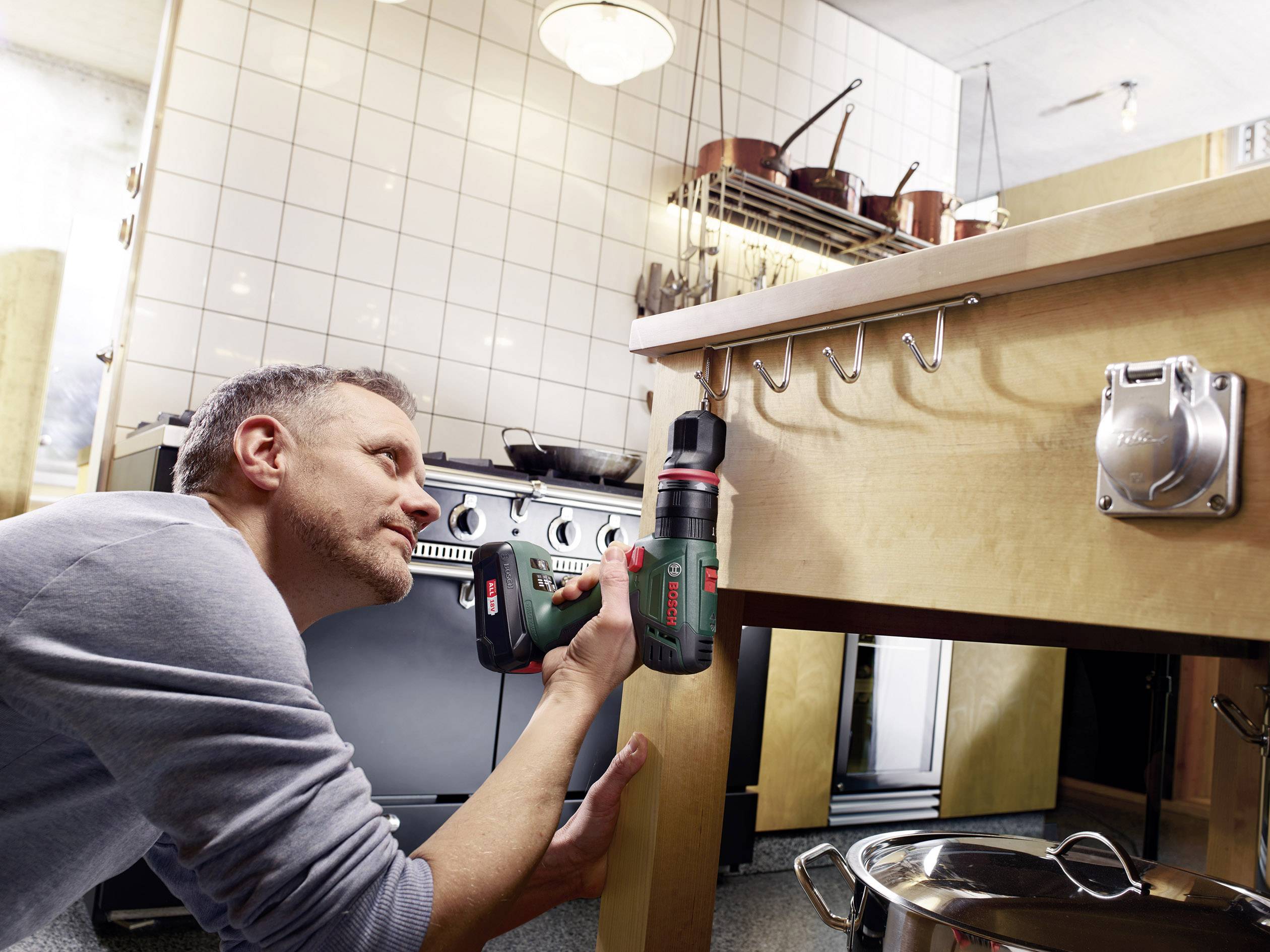 Un homme utilise une visseuse sans fil pour travailler sous un plan de travail de cuisine. Des fourneaux et des meubles suspendus sont visibles en arrière-plan.