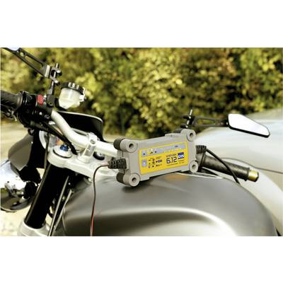 Un chargeur de batterie est fixé sur une motocyclette garée en extérieur. L'appareil affiche une tension de 6,12 volts.
