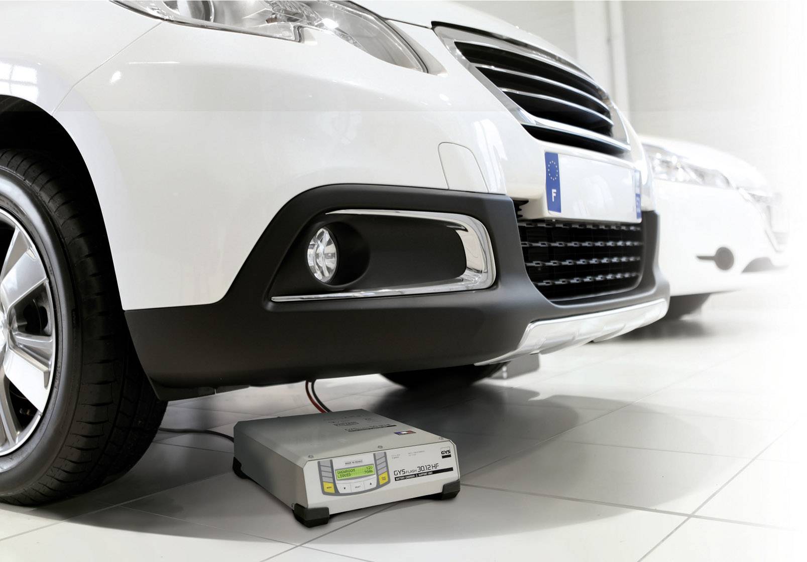 Une voiture blanche avec un chargeur de batterie posé au sol en dessous, relié au véhicule. Elle est stationnée dans une pièce lumineuse.