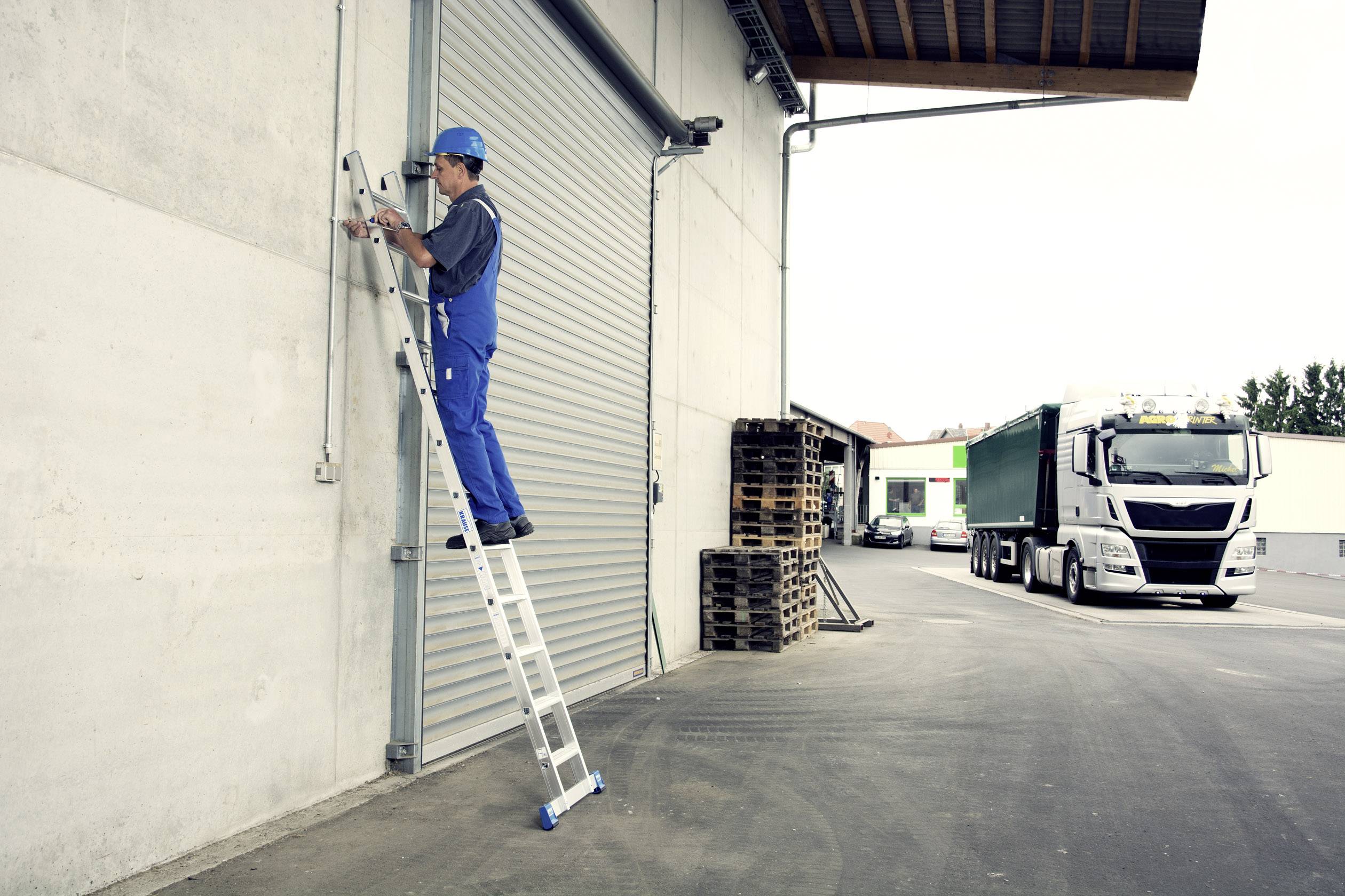 Un ouvrier sur une échelle répare un portail industriel, tandis qu'un camion est stationné en arrière-plan. Des palettes sont empilées contre le mur.
