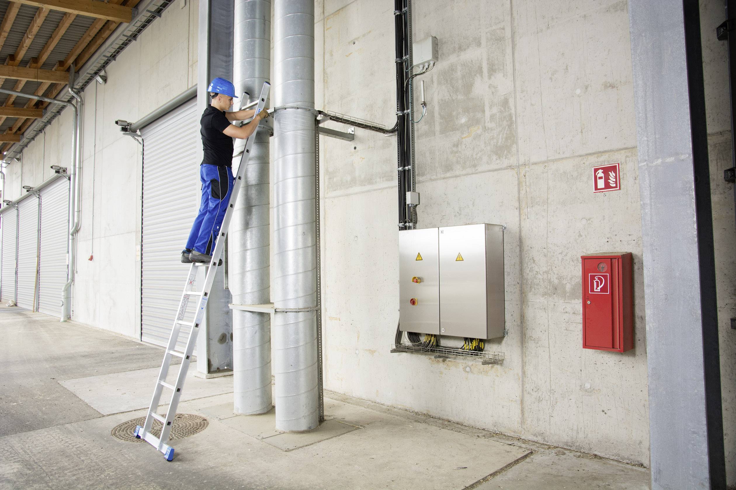 Un ouvrier est debout sur une échelle et travaille sur des tuyaux contre un mur de bâtiment. Un équipement de sécurité, comme un casque, est porté.
