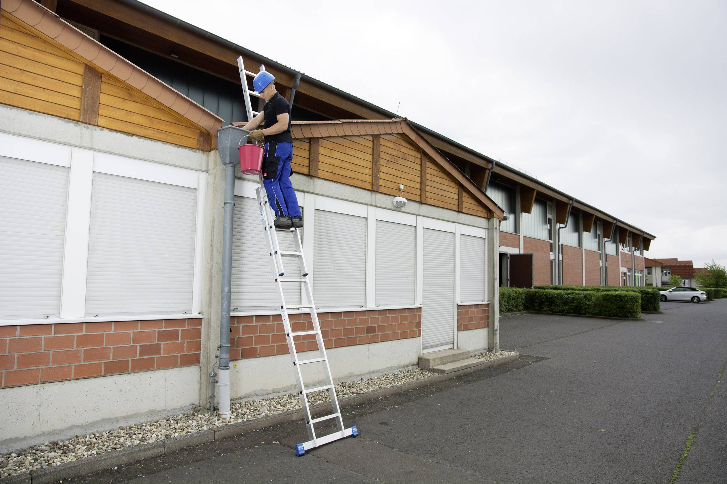 Une personne sur une échelle nettoie une gouttière d'un bâtiment. Elle porte un équipement de sécurité et s'appuie sur le bord de la gouttière.