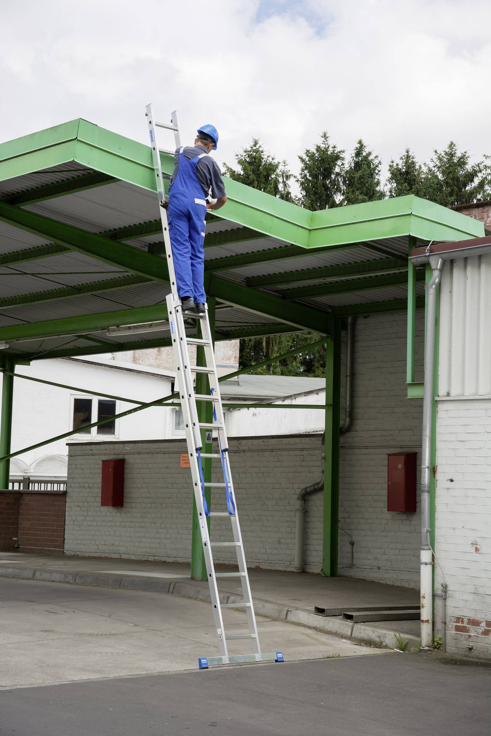 Une personne vêtue de bleu est perchée sur une échelle et travaille sur un bâtiment avec des structures métalliques vertes à l'extérieur.