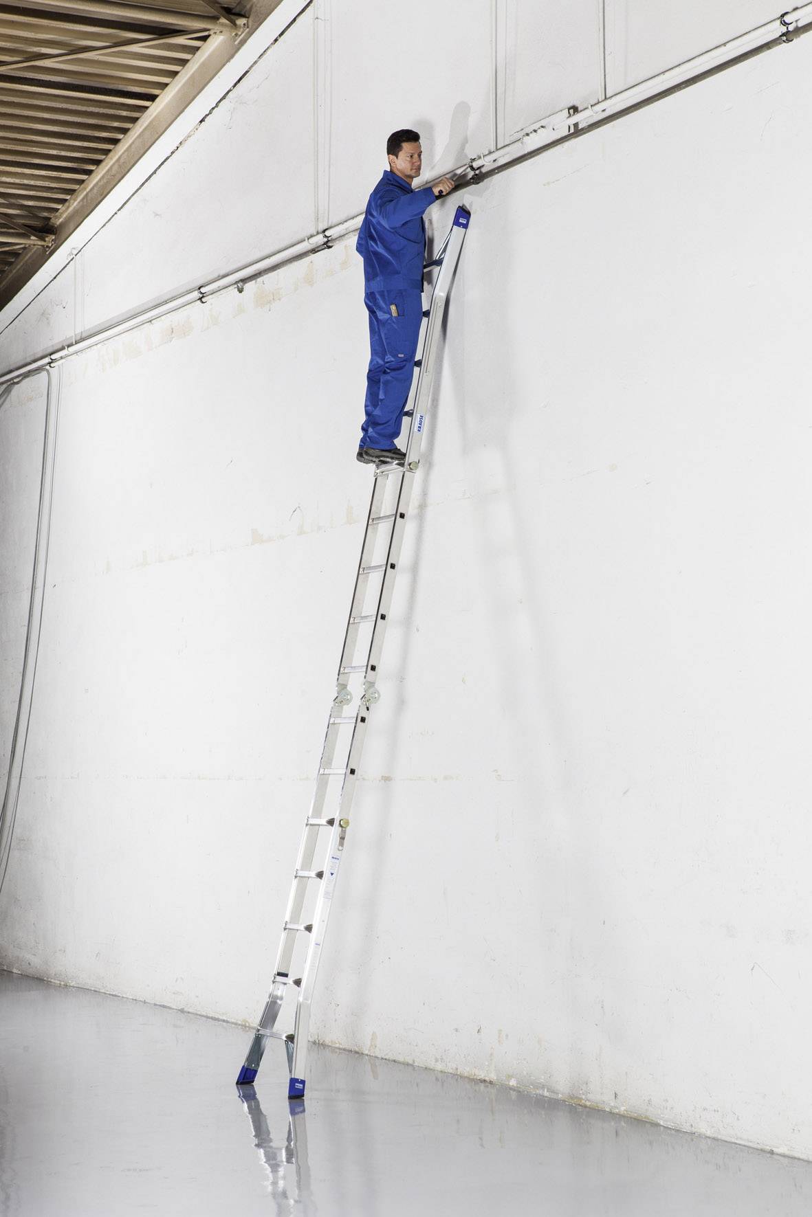 Un homme se tient au sommet d'une longue échelle appuyée contre un mur blanc élevé, dans une grande pièce au sol réfléchissant.