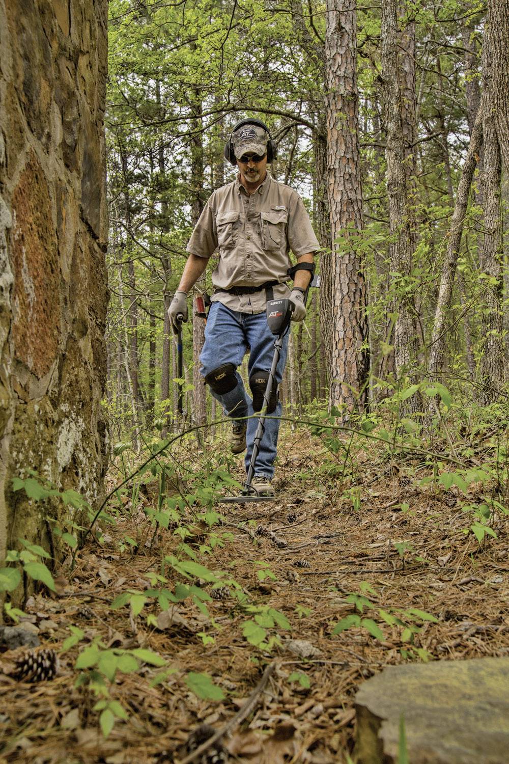 Une personne dans la forêt utilise un détecteur de métaux pour examiner le sol. Elle porte des écouteurs et des gants.