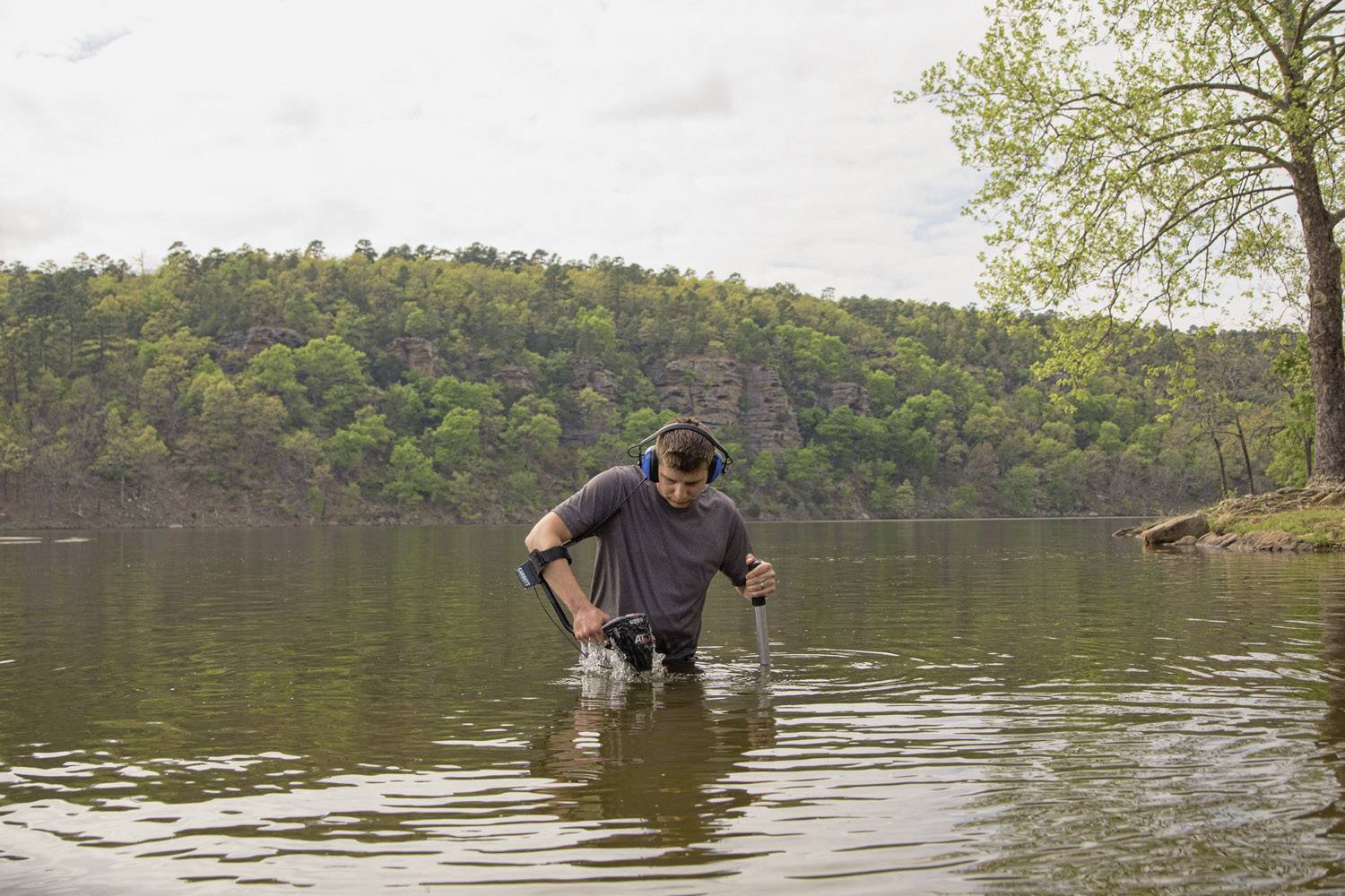 Une personne marche dans l'eau peu profonde d'un lac en utilisant un détecteur de métaux. Des collines boisées sont visibles en arrière-plan.