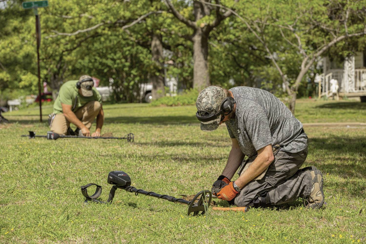 Deux personnes sont à genoux dans un champ et utilisent des détecteurs de métaux pour rechercher des objets métalliques dans le sol. Des arbres et des maisons sont visibles en arrière-plan.