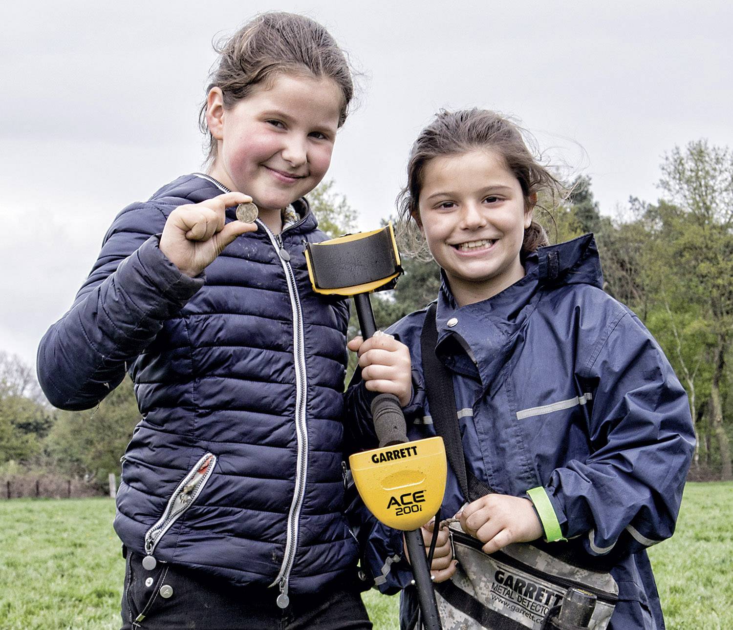 Deux enfants se tiennent joyeusement dans un pré, tenant un détecteur de métaux et montrant fièrement une petite découverte à la caméra.