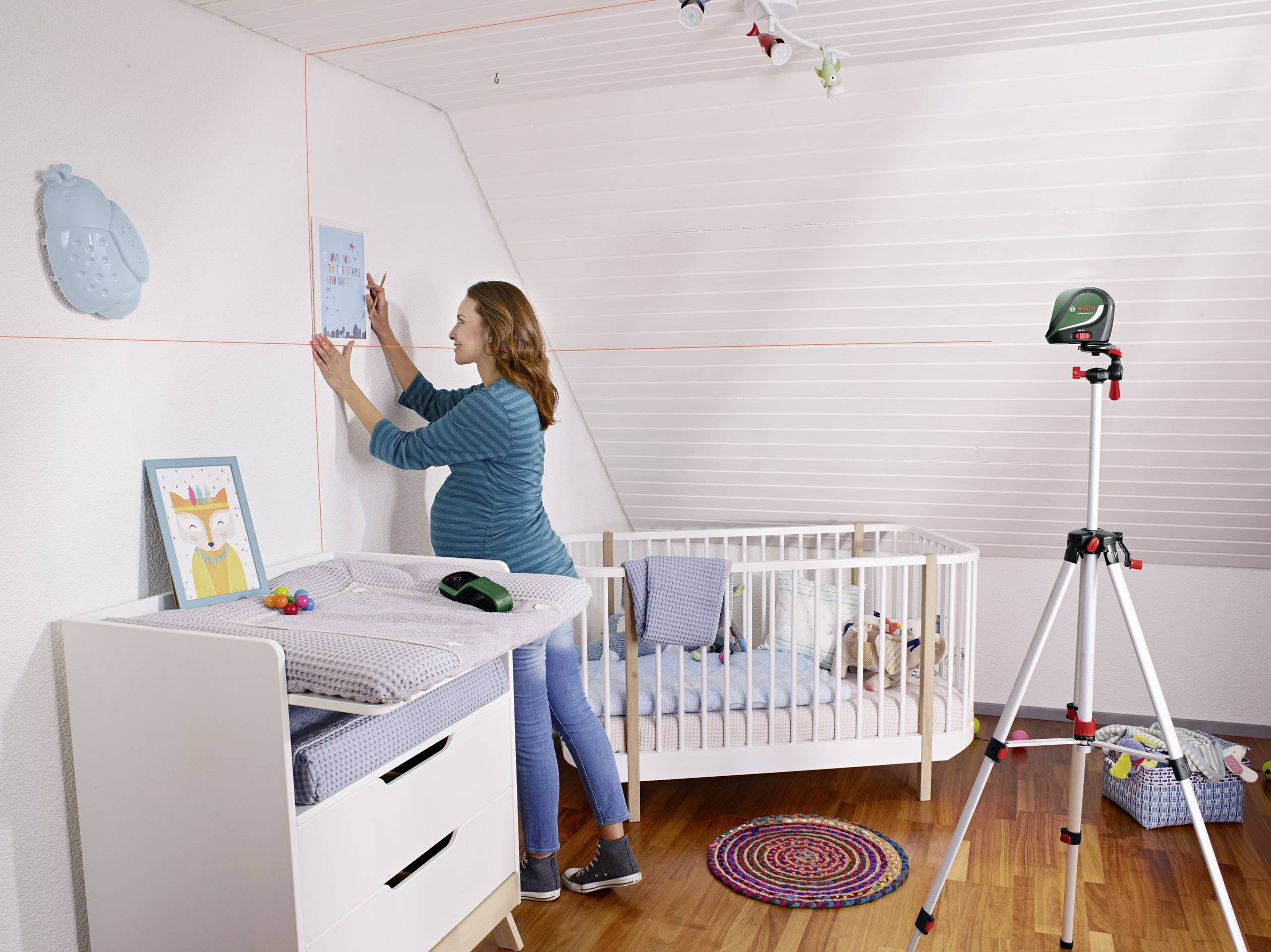 Une femme accroche un tableau dans la chambre d'enfant. Un bébé dort dans un lit à barreaux. Un appareil de mesure laser monté sur trépied est installé dans la pièce.