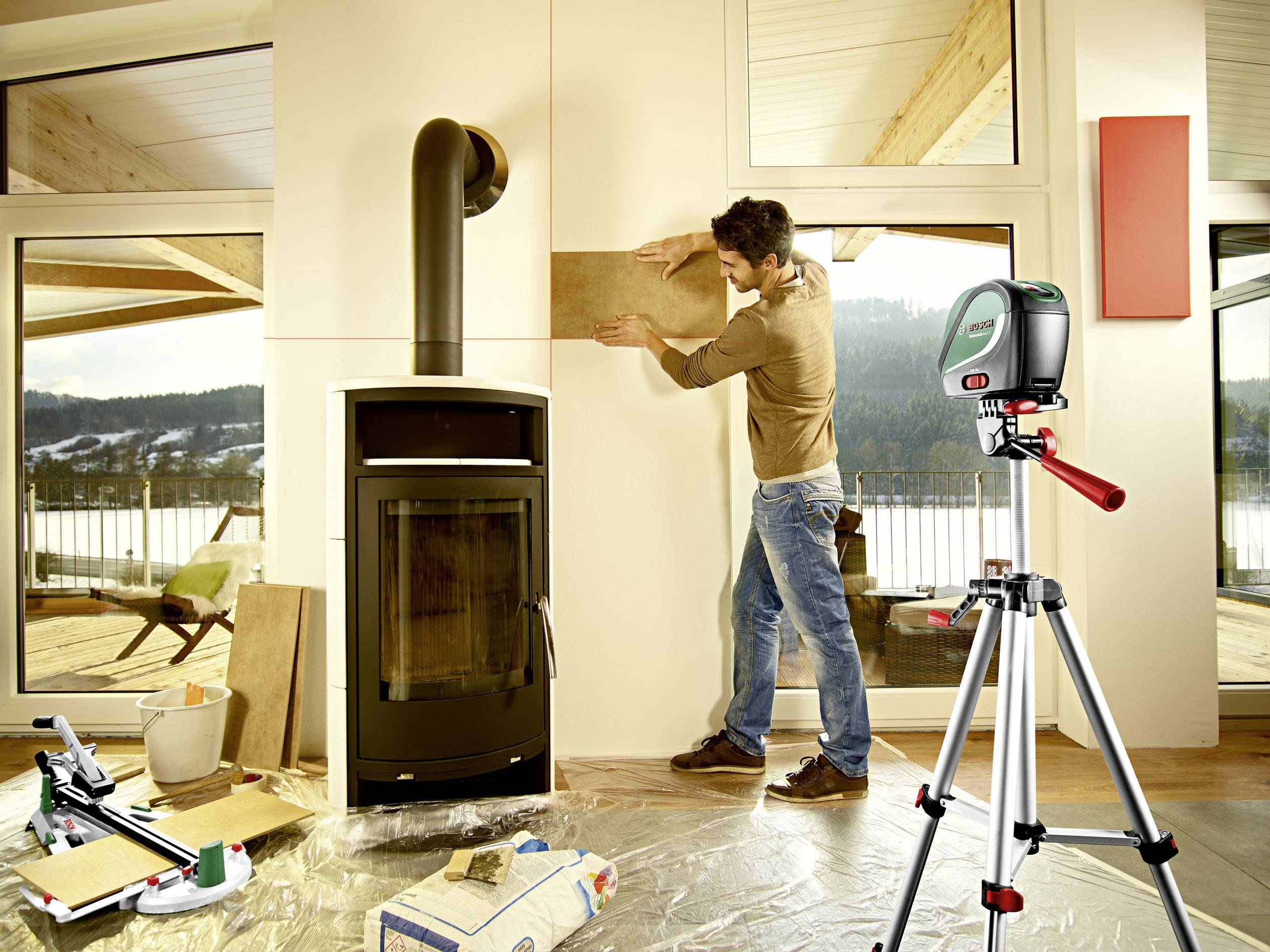 Un homme ajuste une planche de bois sur un mur à côté d'un poêle, assisté par un niveau laser monté sur un trépied.