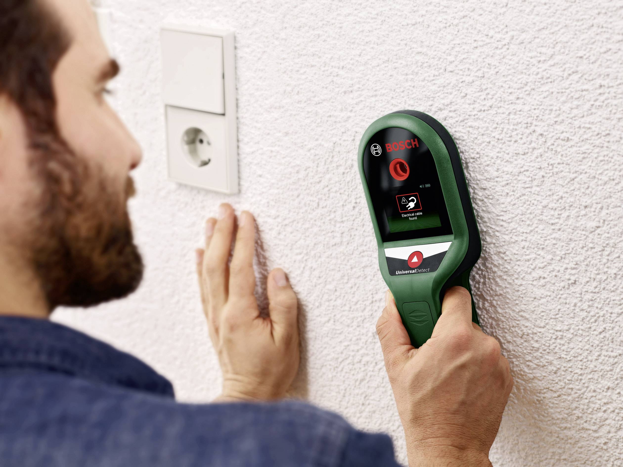 Un homme utilise un détecteur mural Bosch pour rechercher des câbles et des tuyaux dans un mur blanc. Une prise électrique est visible en arrière-plan.