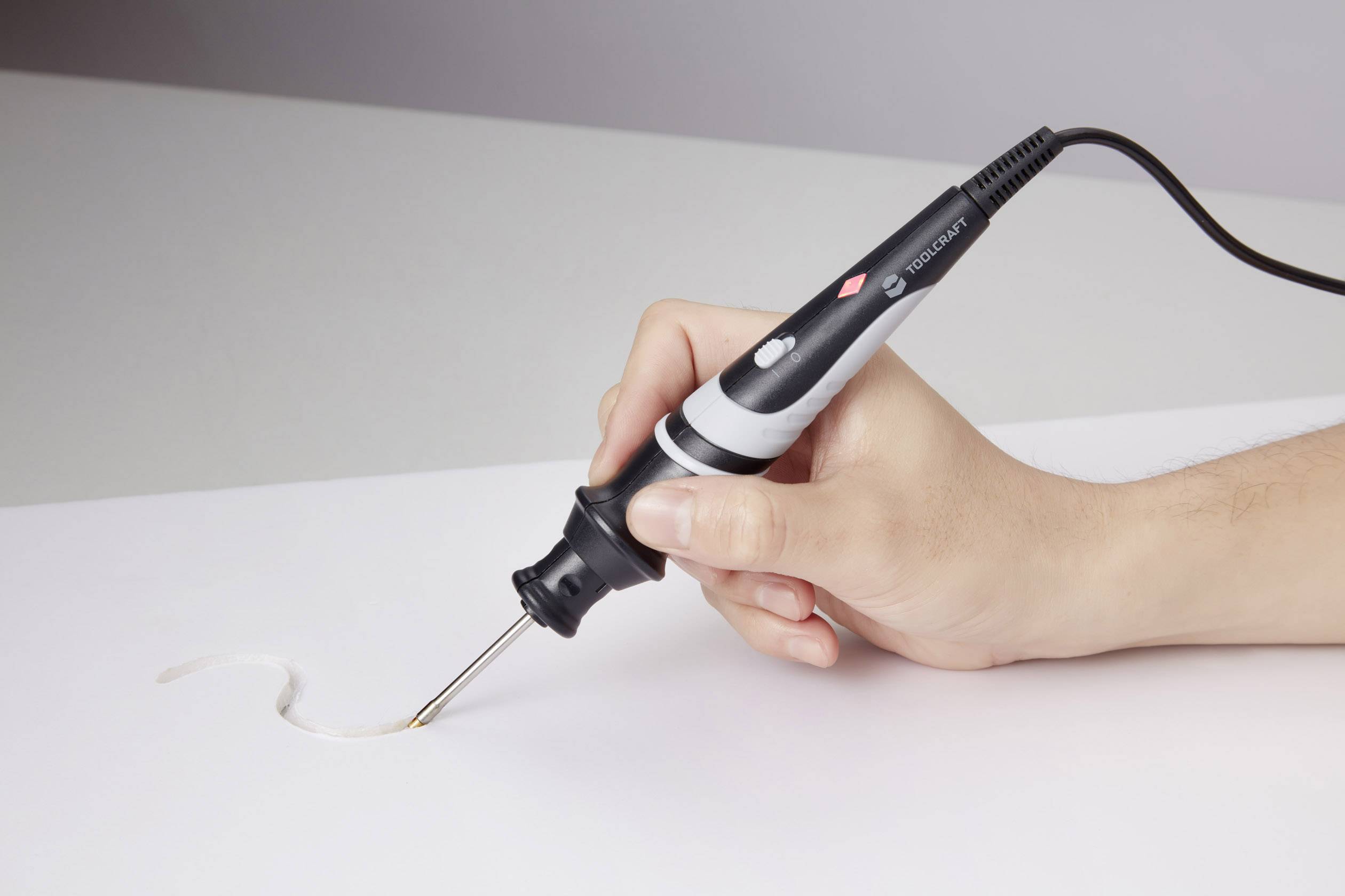 Une main tient un fer à souder dirigé vers une feuille de papier blanche. Le fer à souder présente un design noir et argenté.