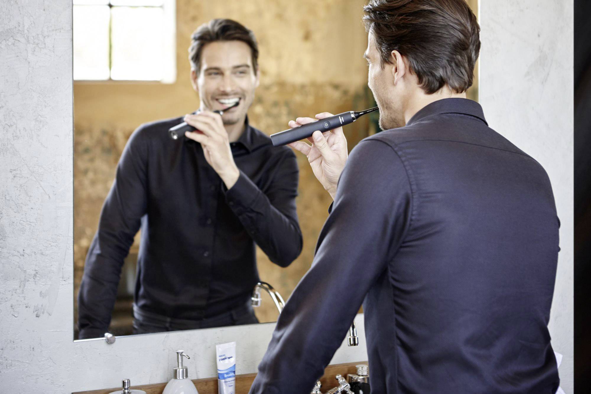 Un homme se brosse les dents en souriant devant un miroir. Il porte une chemise sombre. Un tube de dentifrice et des serviettes sont visibles.