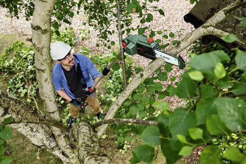 Un homme en vêtements de travail coupe une branche d'arbre avec une tronçonneuse électrique. Il porte un casque de protection et se trouve debout sur le sol.