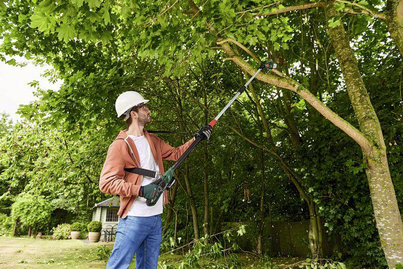 Un homme portant un casque et des lunettes de protection utilise une tronçonneuse fixée à un manche pour élaguer les branches d'un arbre dans un jardin.