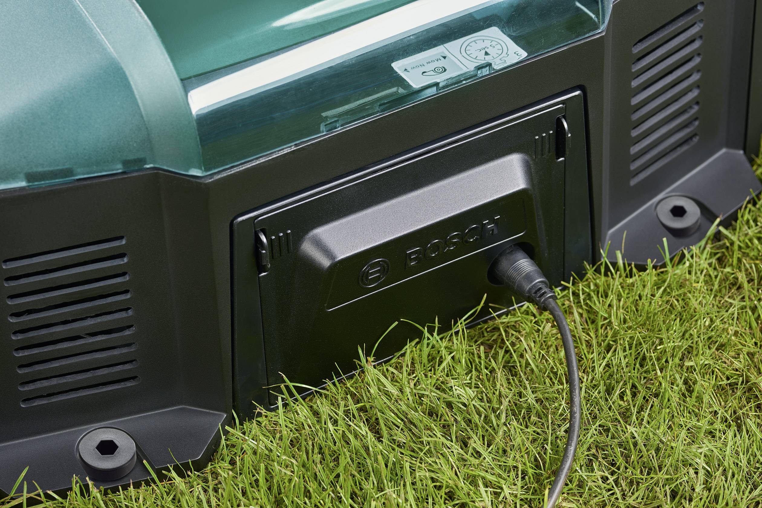 Un robot tondeuse sur sa station de charge, branché par un câble. Entouré d'herbe verte.