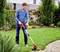 Un homme tond la pelouse avec un coupe-bordures électrique dans un jardin. Des buissons et une maison sont visibles en arrière-plan.