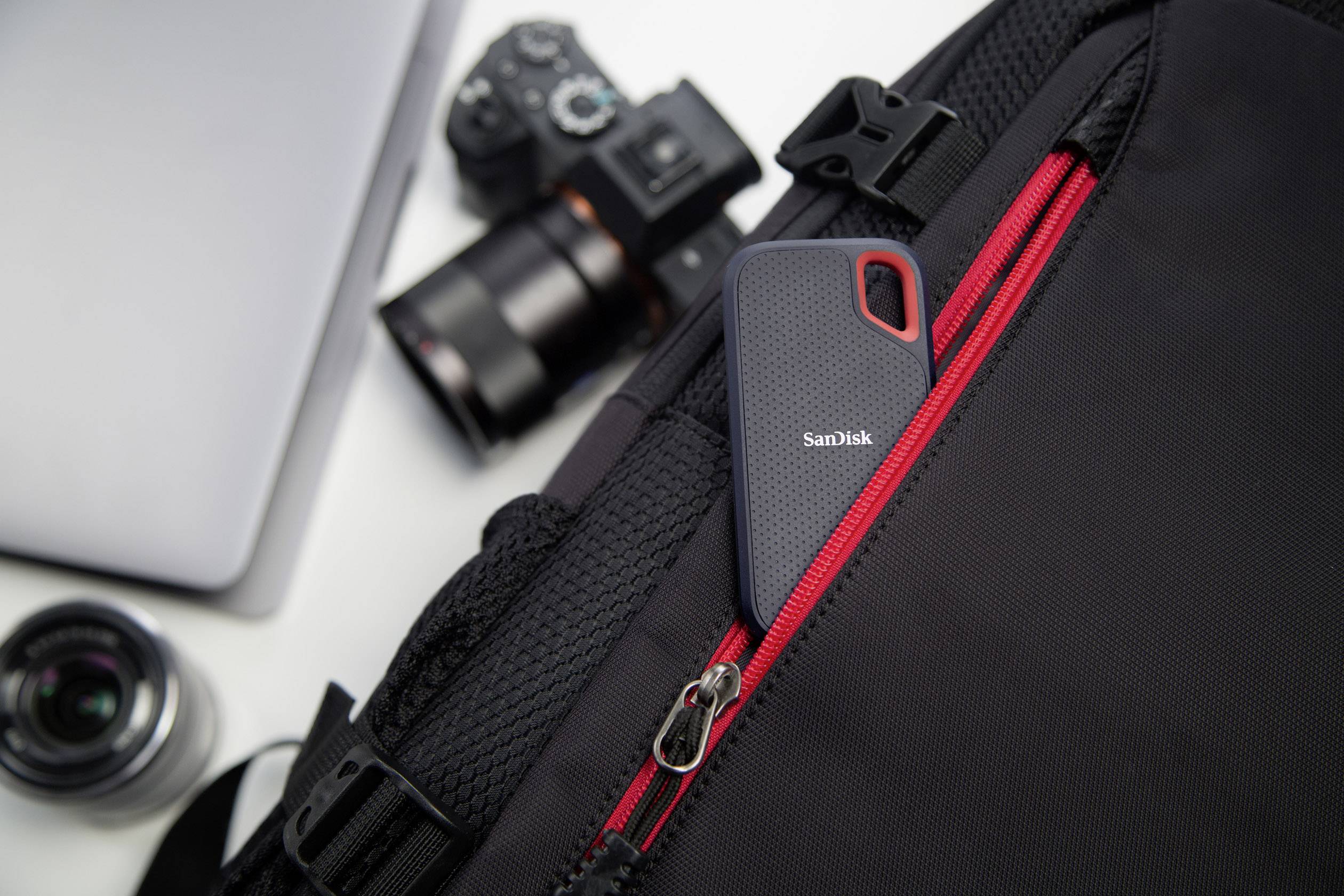 Un disque dur portable SanDisk est glissé dans la poche extérieure d'un sac à dos noir. Un appareil photo et un ordinateur portable sont visibles en arrière-plan.