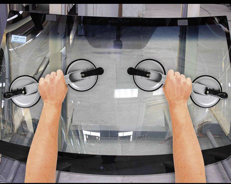 Deux mains tiennent une vitre de voiture avec des ventouses. La scène illustre l'installation ou le remplacement du pare-brise dans un atelier.