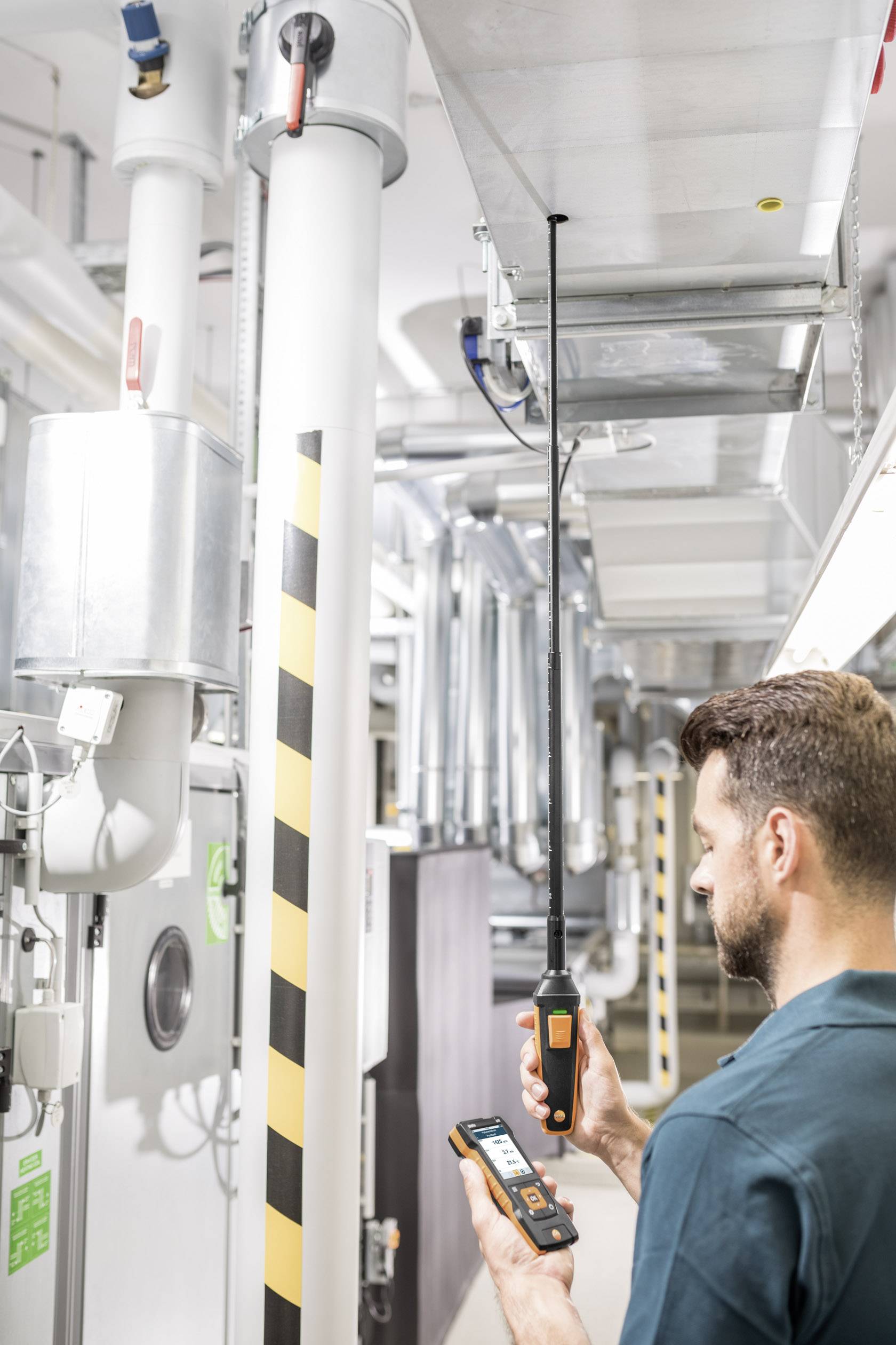 Une personne vérifie les systèmes de ventilation à l'aide d'un appareil portable dans un bâtiment industriel. Des tuyaux et de l'équipement technique sont visibles.