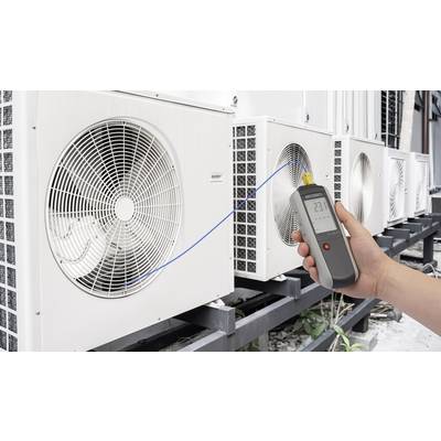 Une main tient un thermomètre numérique devant des unités de climatisation. L'écran affiche '23°C'. L'image démontre la mesure de température.