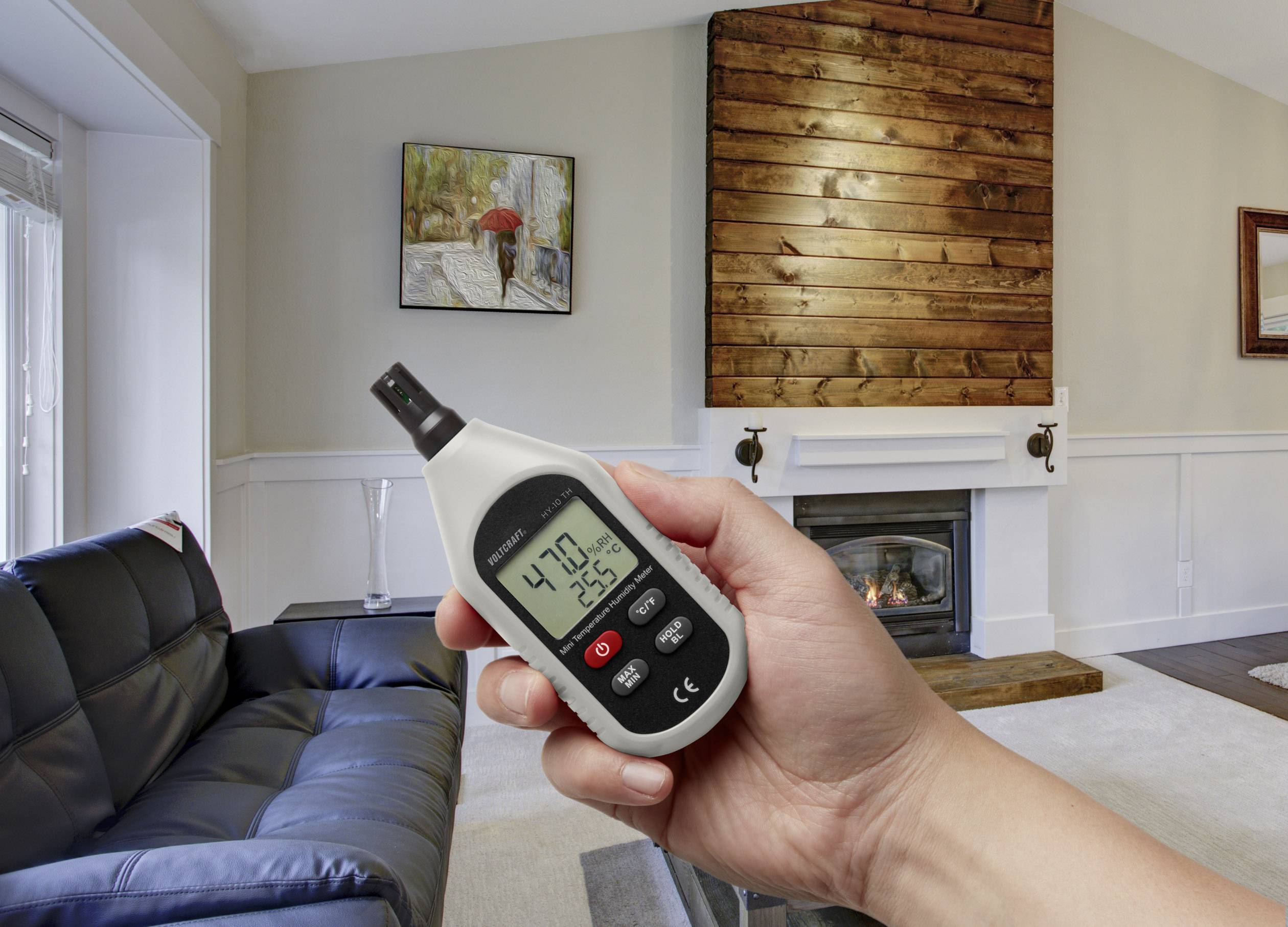 Une main tient un hygromètre dans un salon, affichant 47,0 % d'humidité et 25,6 °C, avec en arrière-plan une cheminée et un canapé.