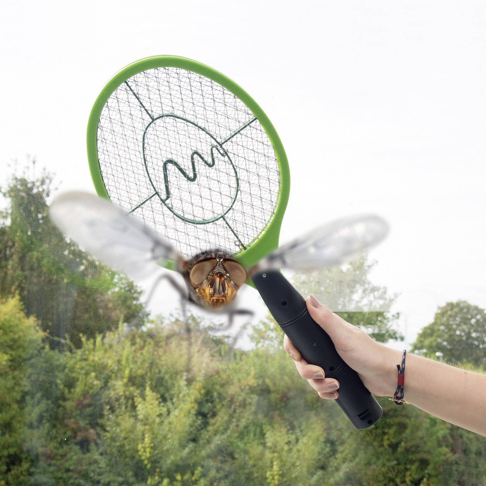 Une main tient un destructeur électrique d'insectes qui vient de frapper une mouche en plein vol. Des arbres flous sont visibles en arrière-plan.