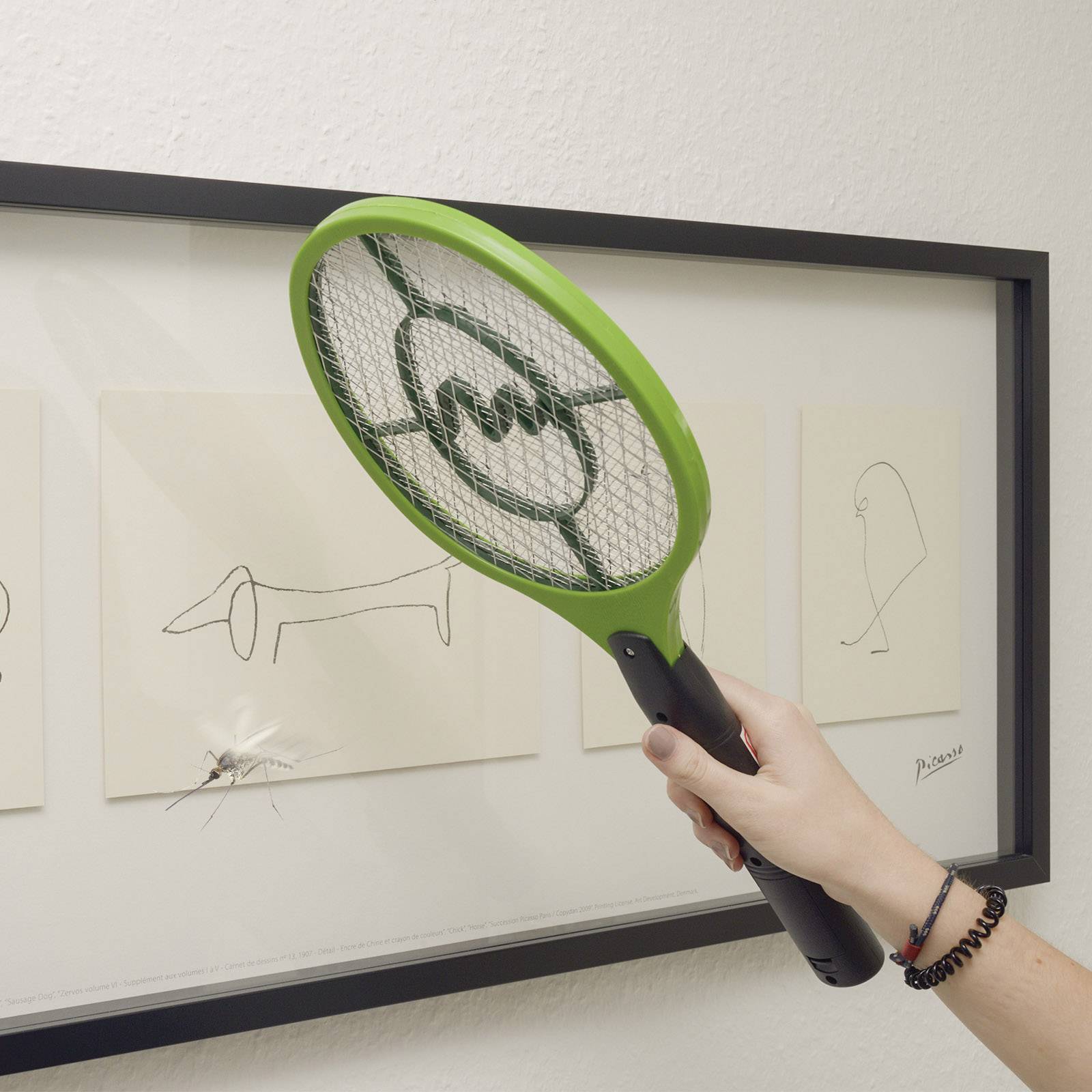 Une main tient une tapette électrique devant une œuvre d'art, tandis que la tapette écrase une mouche posée sur un tableau.