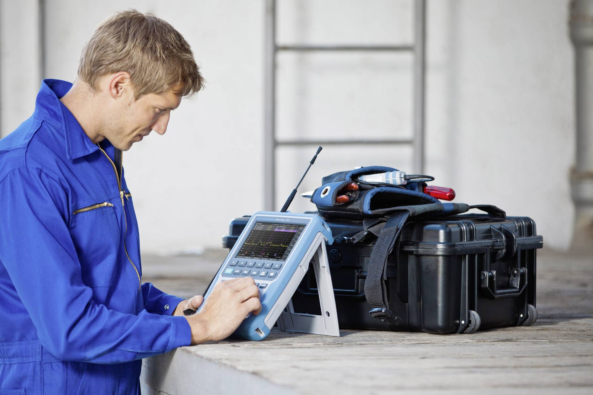 Un technicien en tenue de travail bleue utilise un appareil portable devant une mallette à outils.