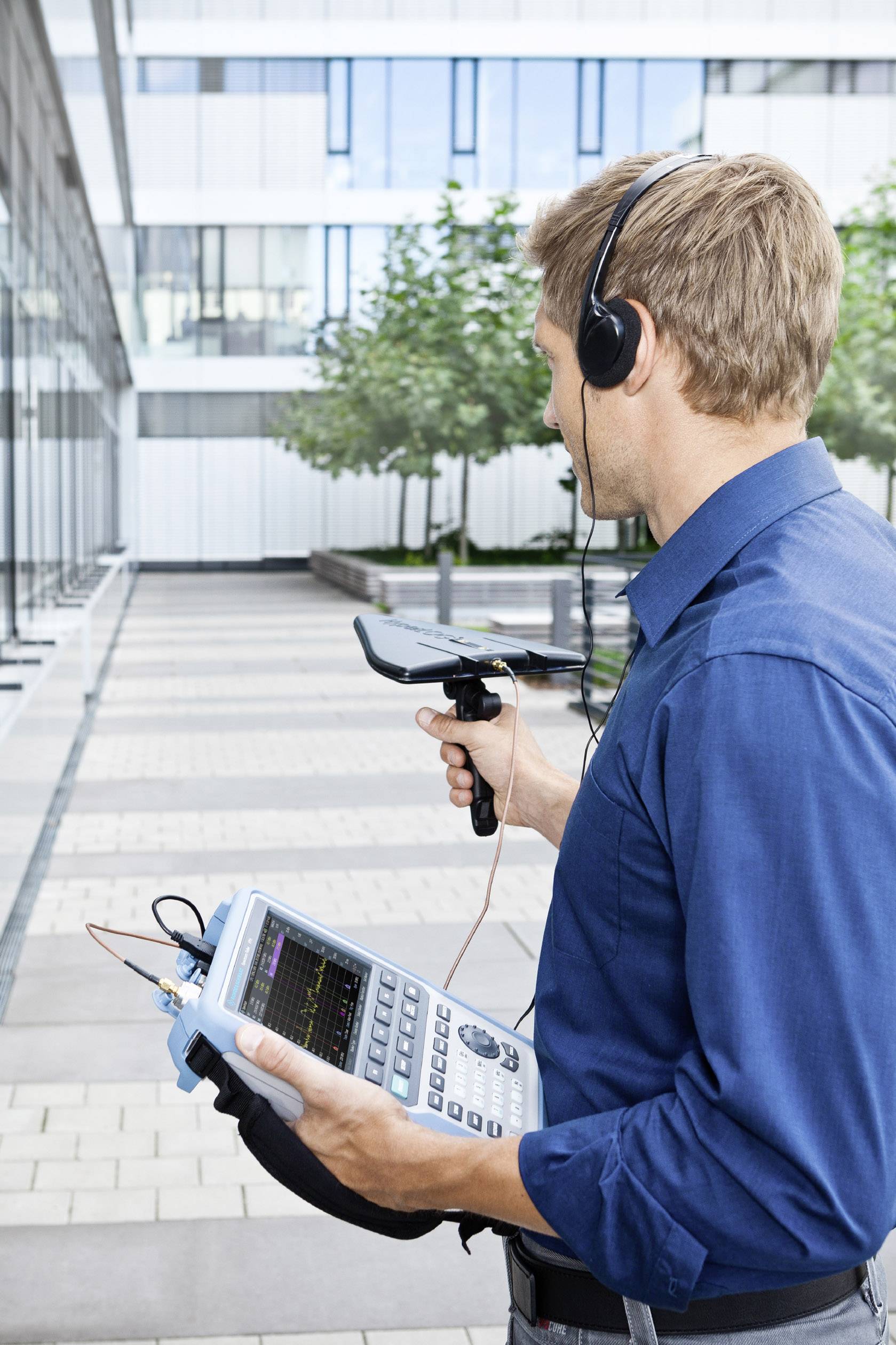 Un homme vêtu de bleu utilise un appareil portable de mesure de fréquence à l'extérieur. Il porte des écouteurs et tient une antenne.
