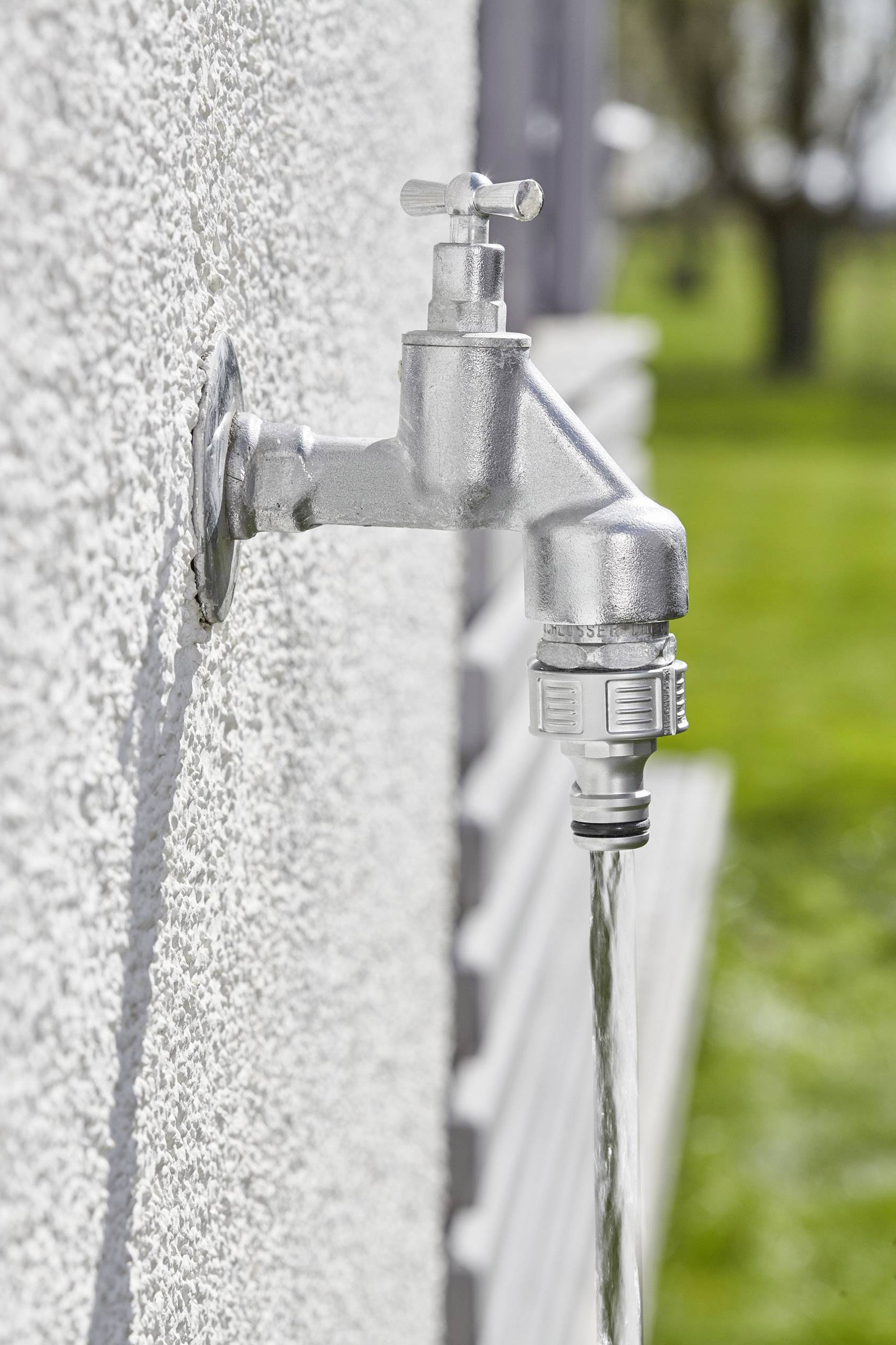 Un robinet sur un mur blanc laisse couler de l'eau. L'arrière-plan montre une zone de jardin floue en plein jour.