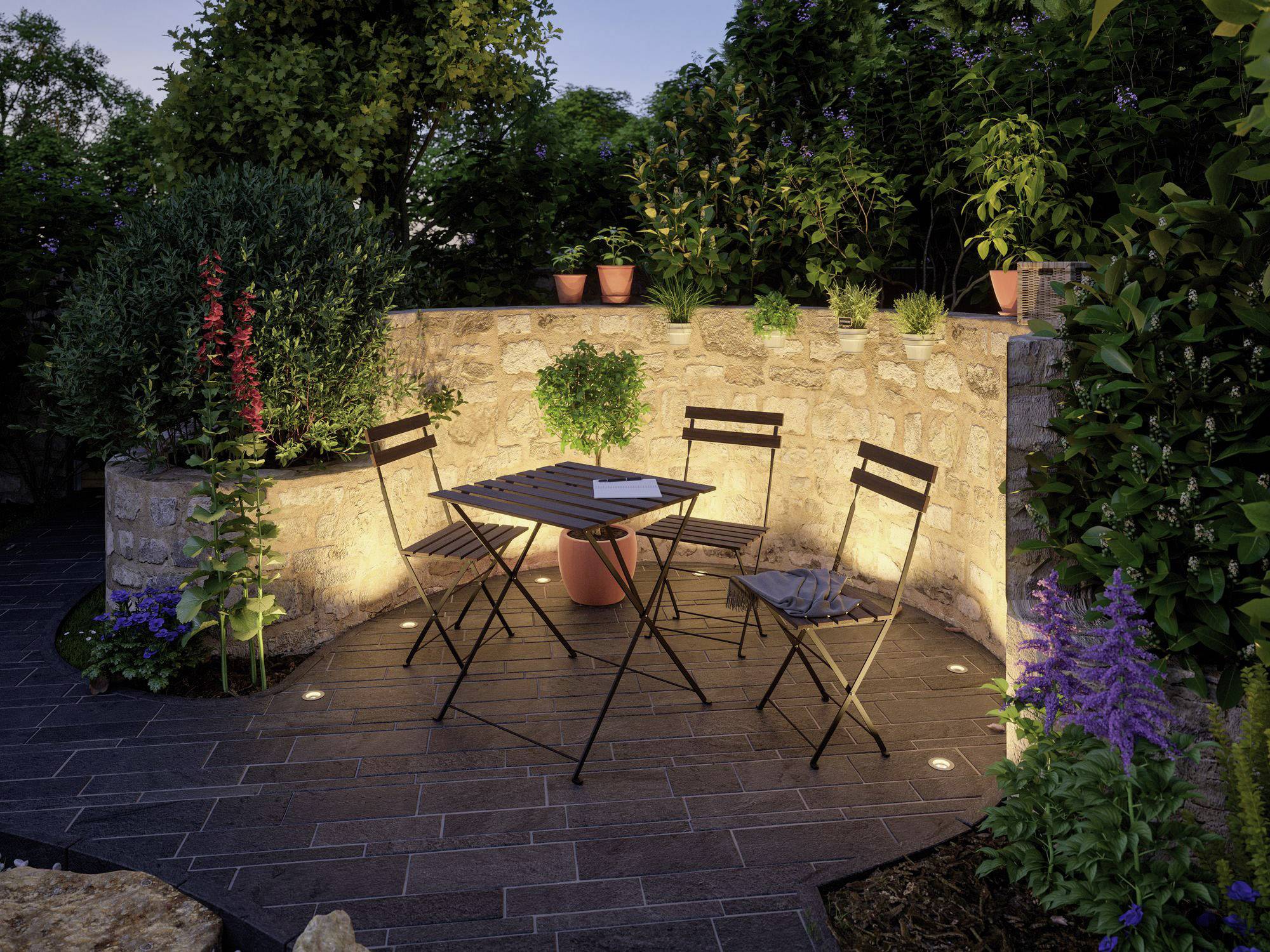 Un petit espace de jardin au crépuscule avec une table ronde et quatre chaises, entouré de plantes et de murs de pierre éclairés.