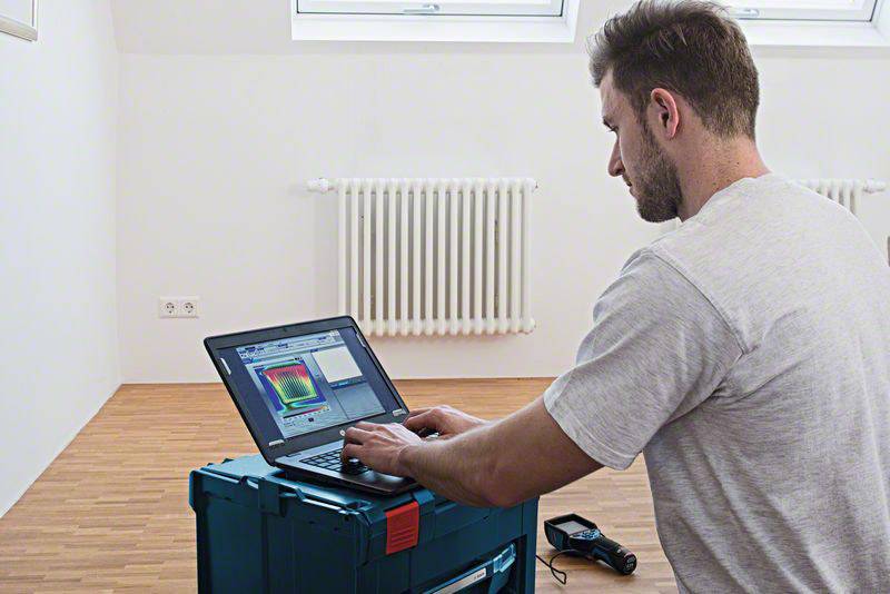 Un homme travaille sur l'image thermique d'une pièce avec un ordinateur portable. Un radiateur est visible en arrière-plan.