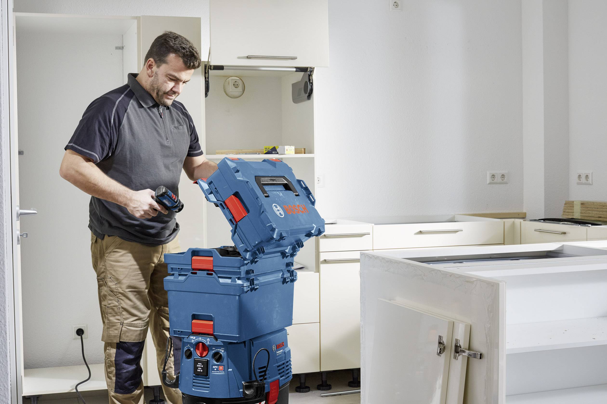 Un homme souriant, vêtu de vêtements de travail, utilise un aspirateur bleu dans une cuisine tout en ouvrant un récipient sans outil.