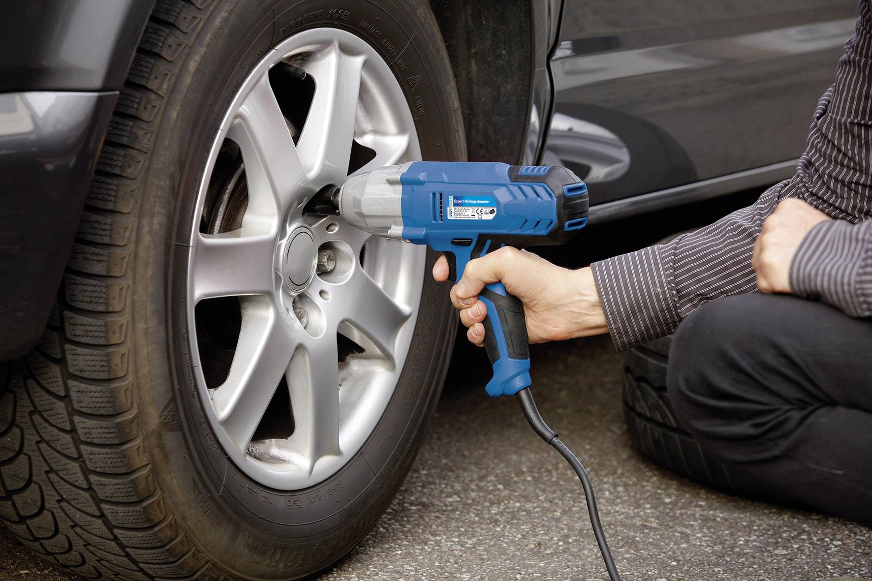 Une personne utilise un tournevis électrique bleu pour changer une roue d'une voiture sur une route pavée.