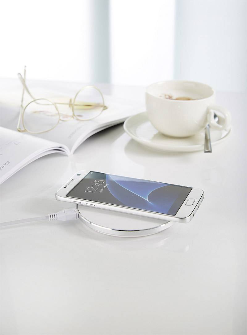 Un smartphone charge sans fil sur une station de charge. À côté se trouvent des lunettes, un magazine ouvert et une tasse de café.