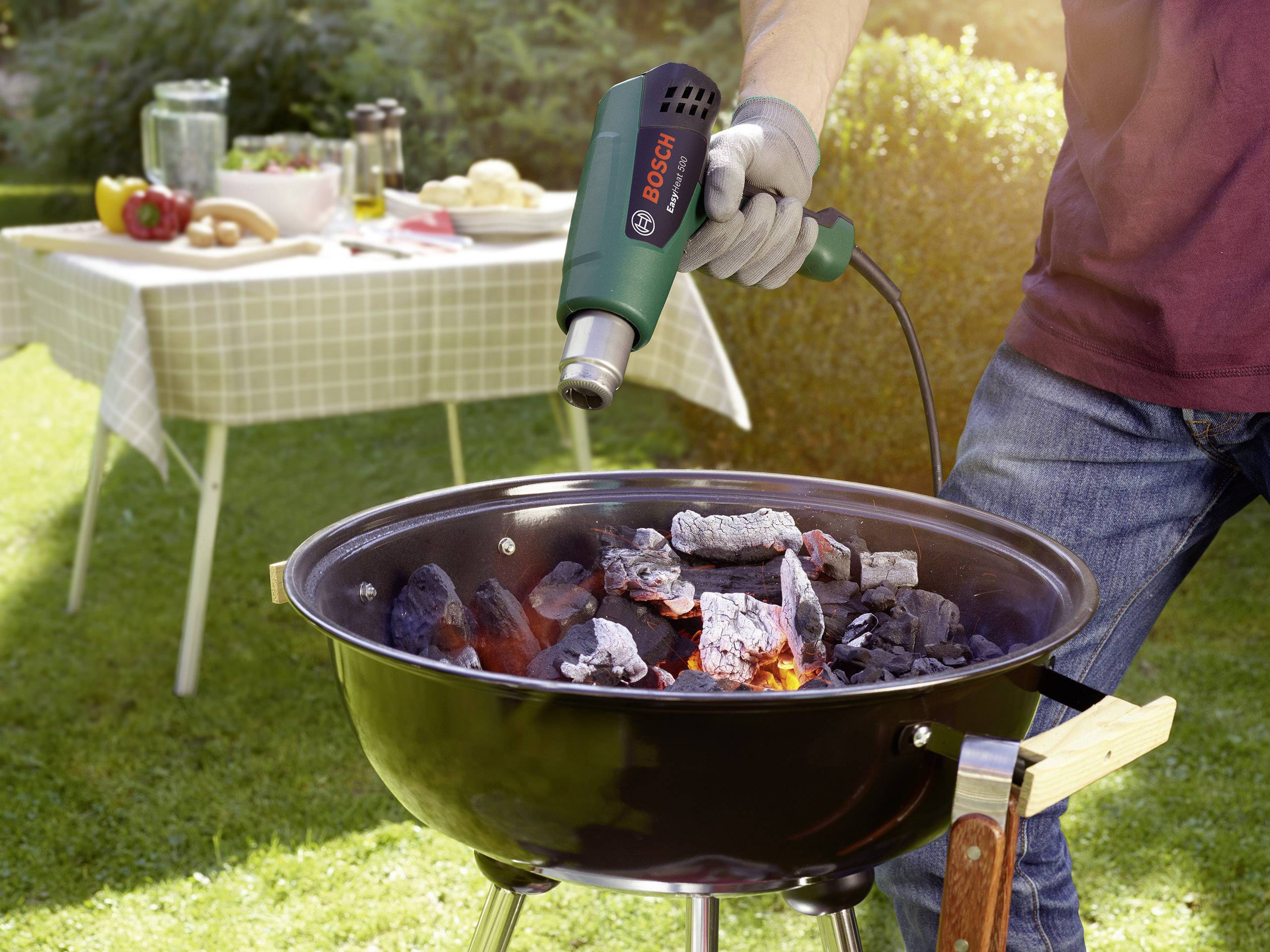 Une personne chauffe du charbon de gril avec un souffleur d'air chaud dans le jardin. À l'arrière-plan, une table est dressée avec de la nourriture et des boissons.