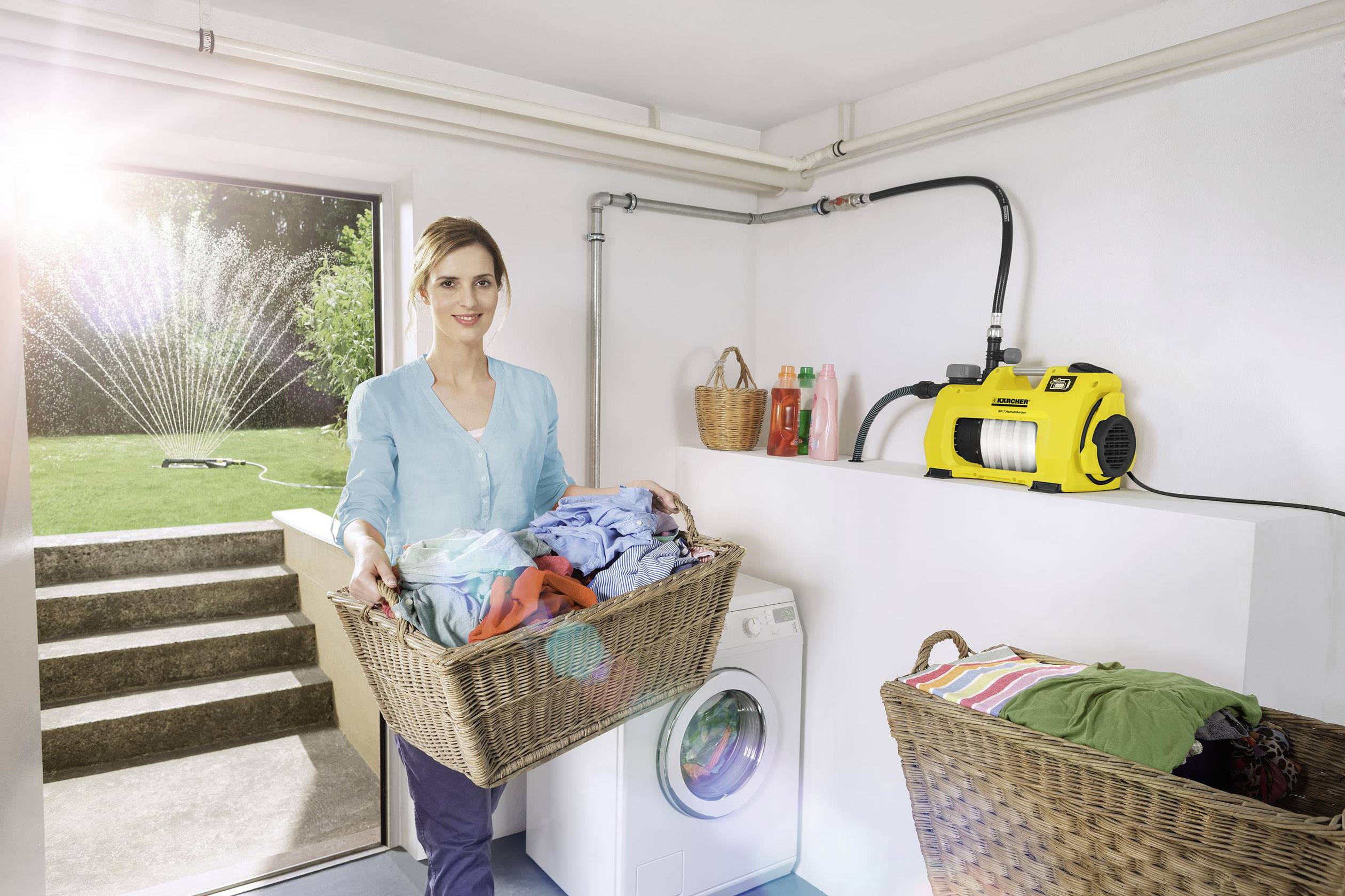 Une femme se tient dans la buanderie avec un panier à linge, un arroseur arrose le jardin en arrière-plan. Une pompe jaune est fixée au mur.