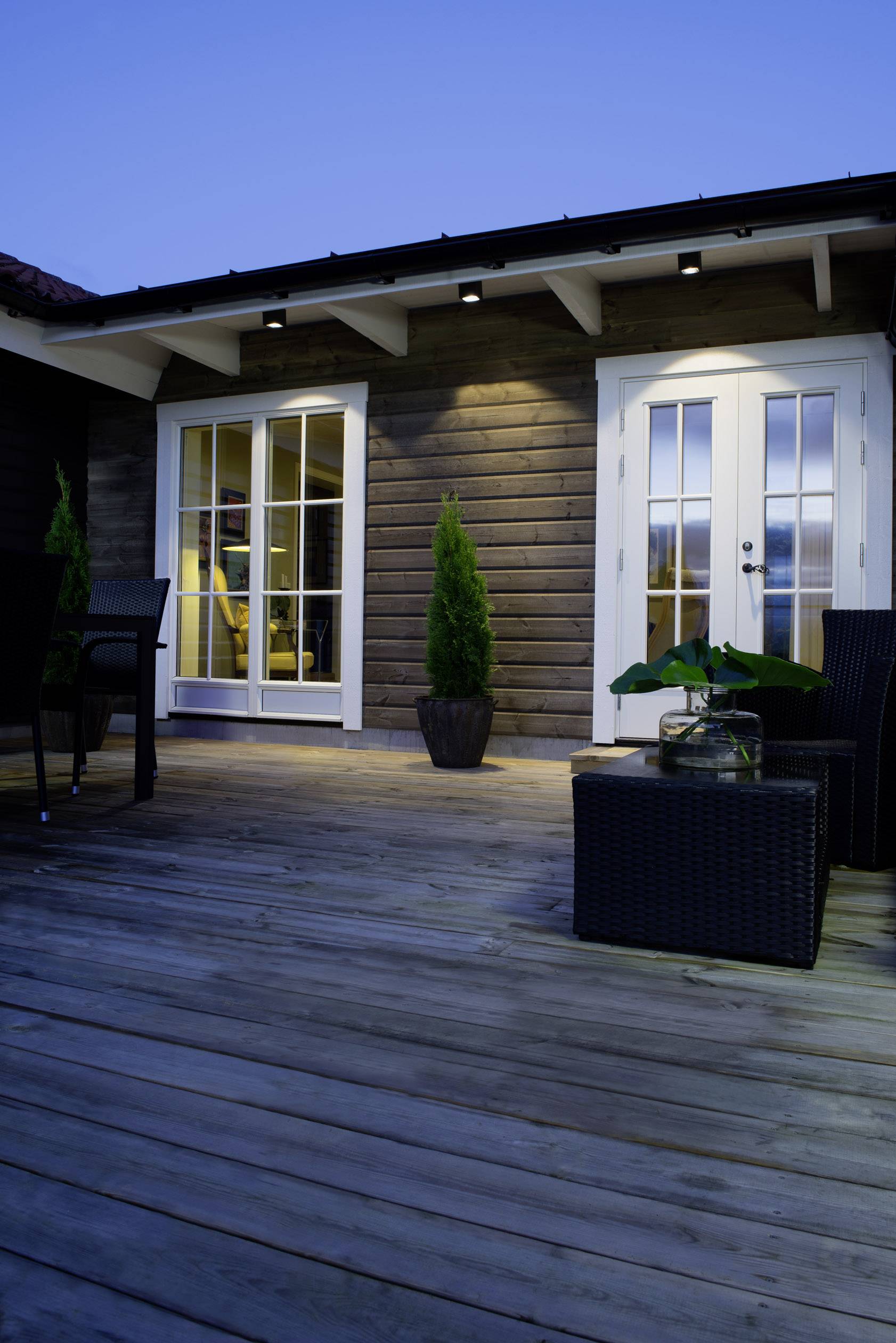 Terrasse en bois au crépuscule avec un ensemble de meubles de jardin noir, deux portes vitrées donnant sur un intérieur éclairé et de petites plantes.
