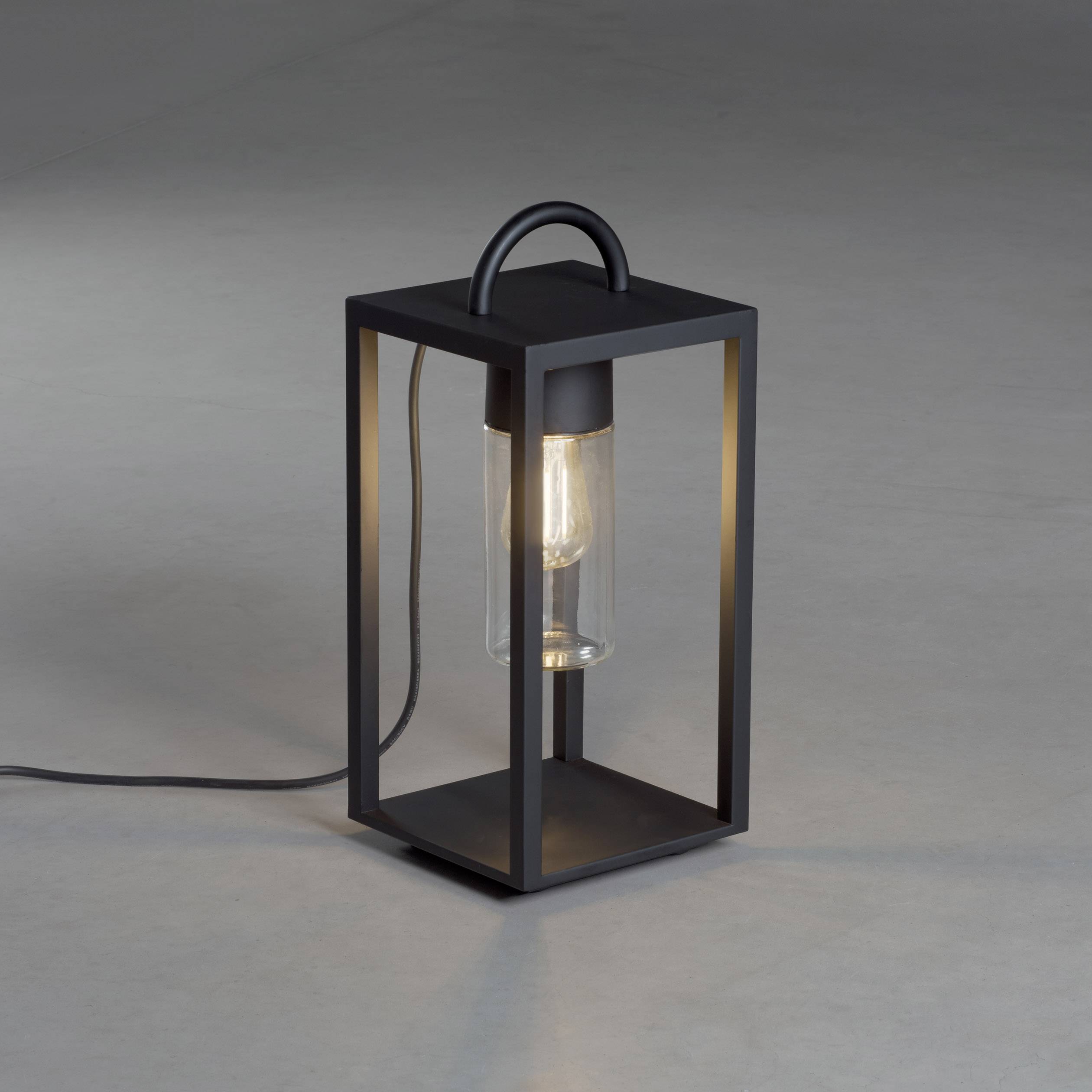 Une lanterne moderne noir avec un boîtier en verre et un éclairage visible est posée sur un sol gris. Le câble longe le sol.