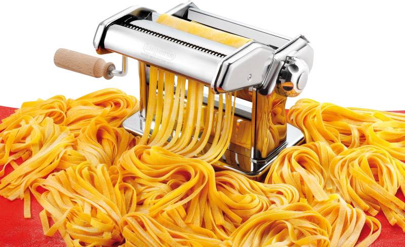 Une machine à pâtes sur une table rouge prépare des pâtes fraîches en ruban. La machine est en métal avec une poignée en bois, formant des pâtes de différentes longueurs.