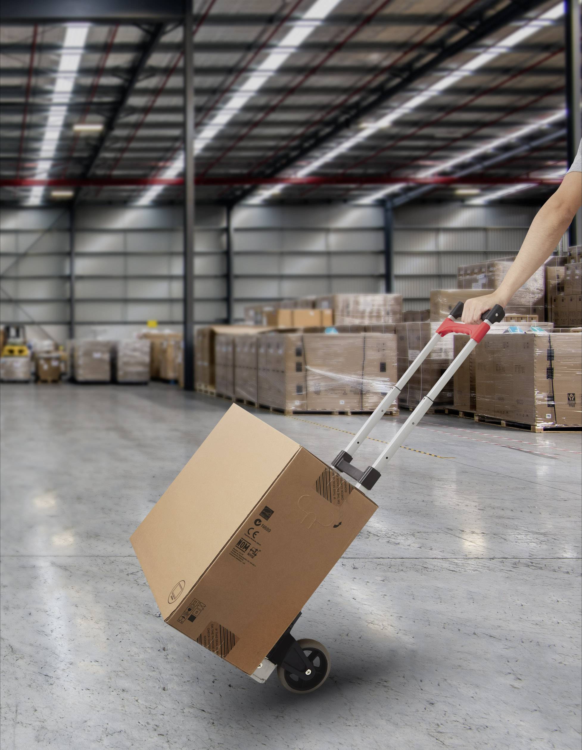 Une personne transporte un grand carton à l'aide d'un diable dans un entrepôt avec des palettes empilées en arrière-plan.
