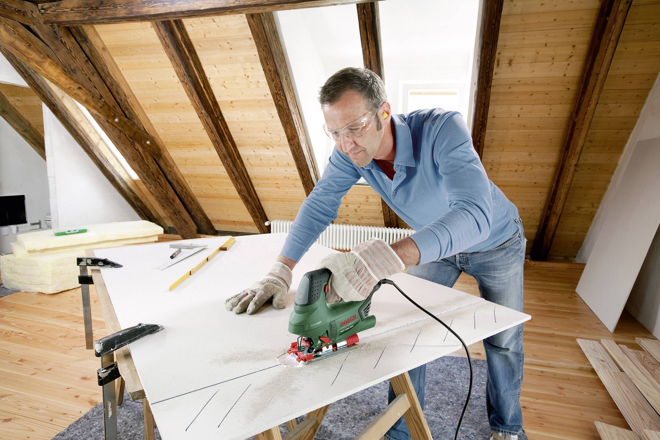 Un homme coupe du contreplaqué avec une scie sauteuse électrique dans un atelier. Il porte des lunettes de sécurité et une protection auditive.