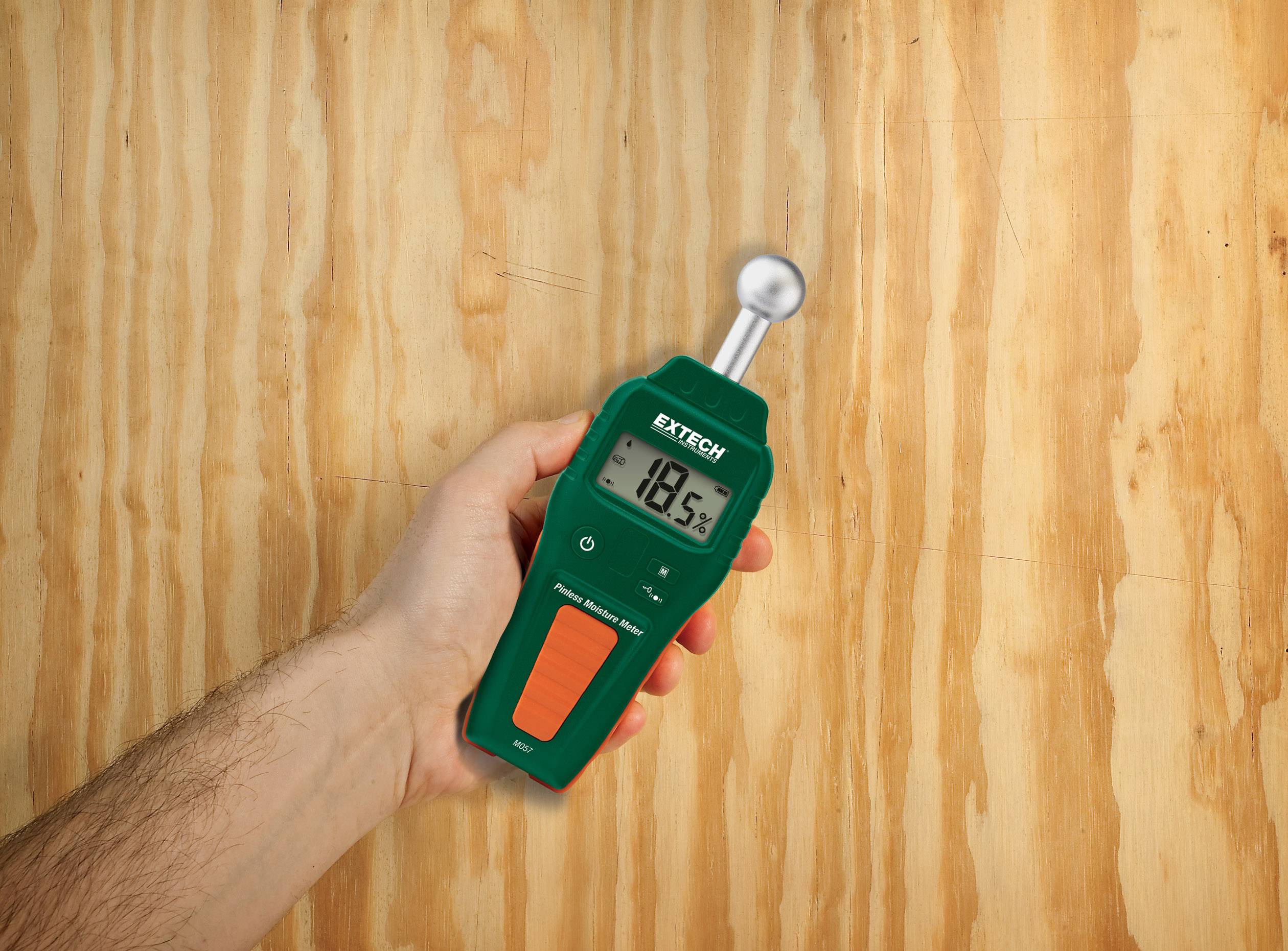 Une main tient un appareil de mesure d'humidité vert affichant 18,5%. Arrière-plan : texture de bois.