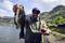Un homme tient un grand poisson au bord d'un lac et sourit à la caméra. Il porte des lunettes de soleil et une casquette.