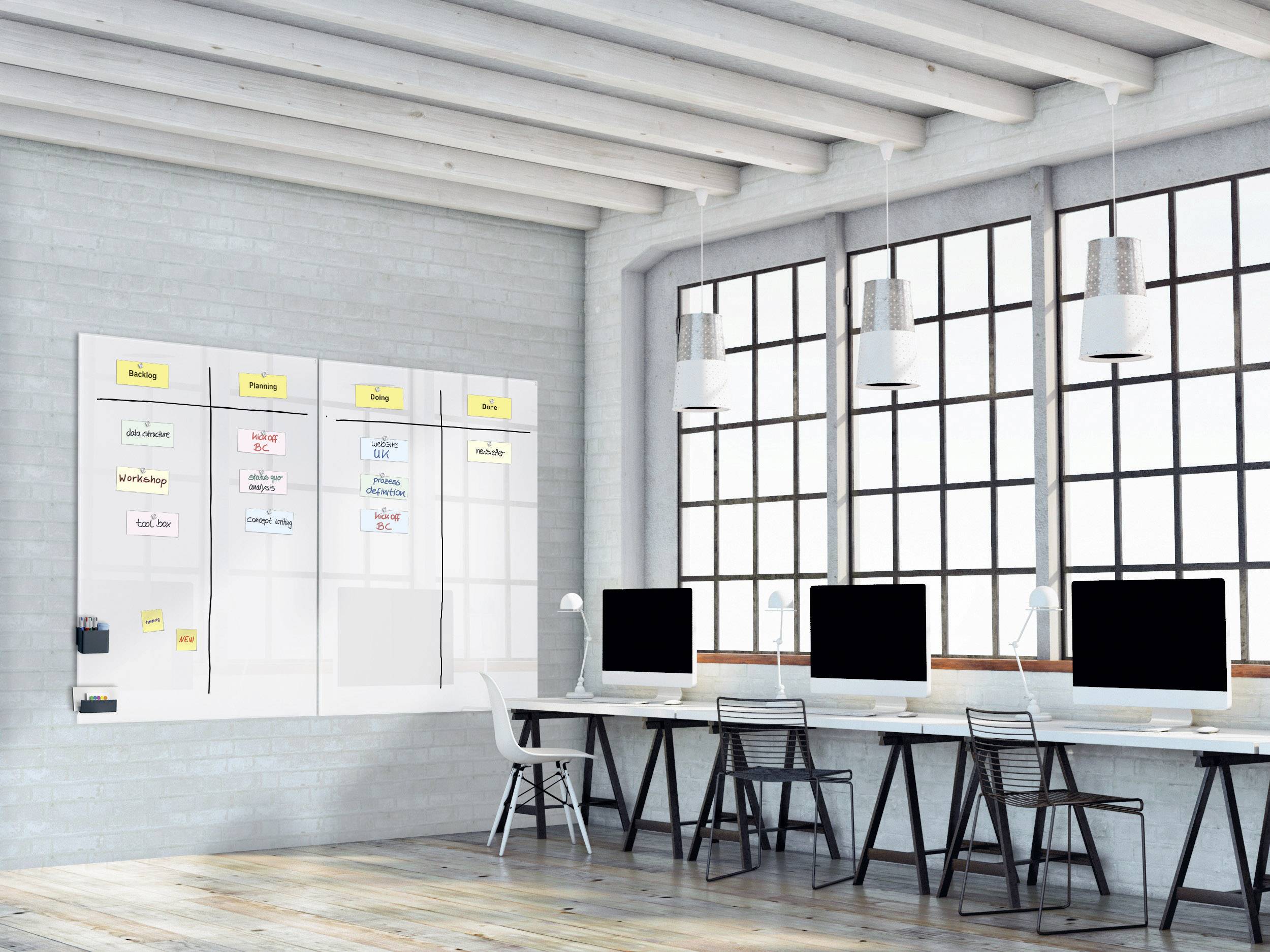 Bureau lumineux avec cinq ordinateurs sur une même table. Sur le mur, un tableau blanc couvert de notes illustrant un processus de travail.