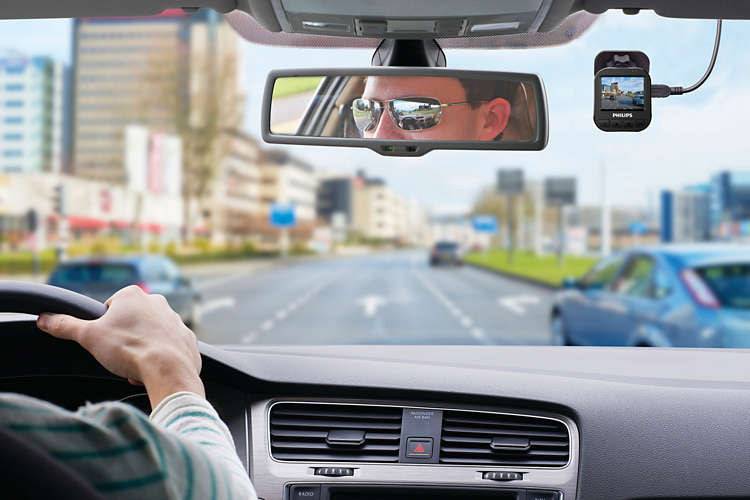 Une personne conduit une voiture dans une ville, avec une dashcam fixée au rétroviseur. La circulation est modérée, avec des bâtiments en arrière-plan.