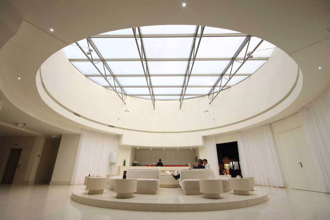 Grand espace lumineux avec un plafond circulaire en verre, en dessous se trouvent des meubles blancs modernes et quelques personnes ; une bar est visible en arrière-plan.
